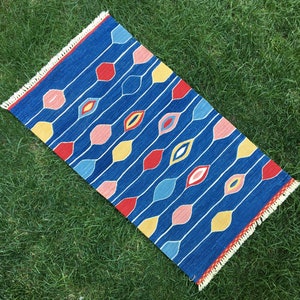 Puede incluir: Alfombra azul con un patrón geométrico de líneas onduladas y óvalos coloridos. La alfombra tiene un borde de flecos.