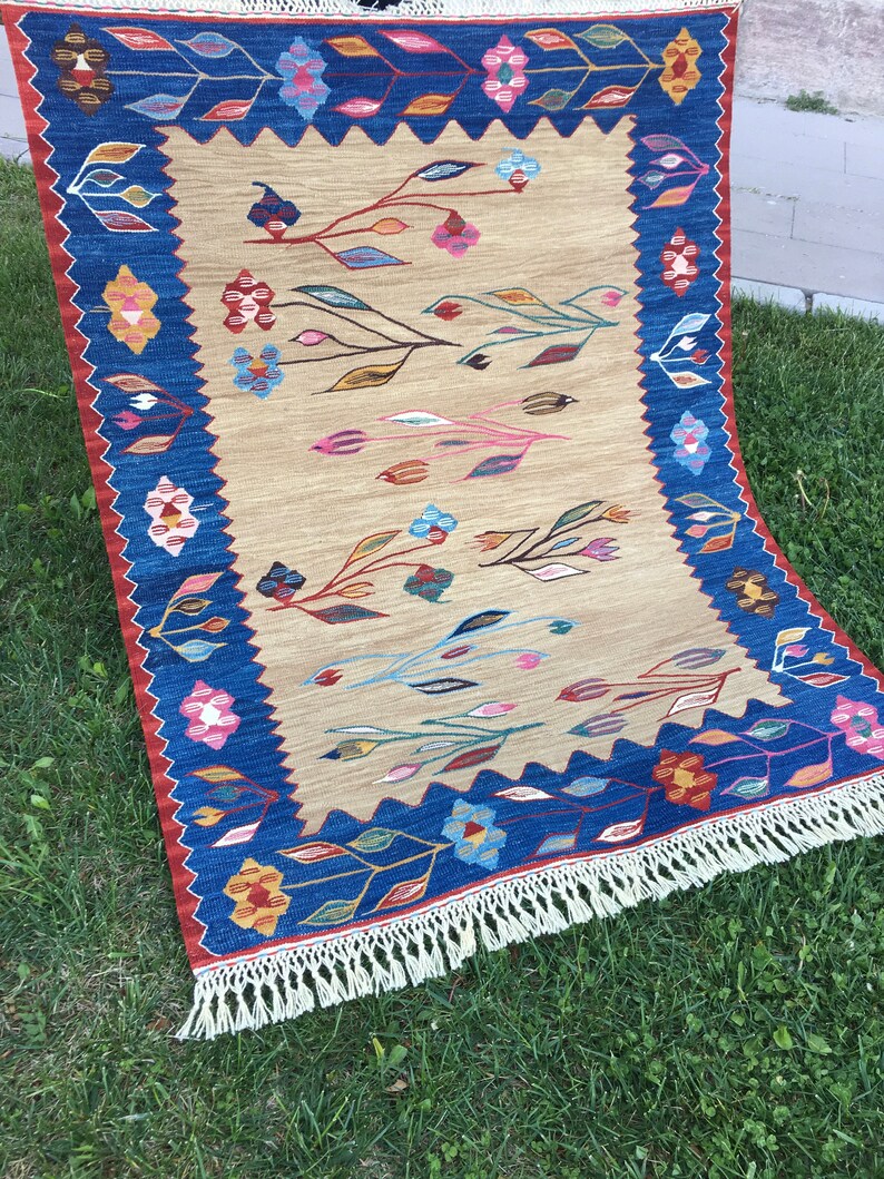 May include: A beige rug with a blue border and a floral pattern. The rug has a fringe on all sides.