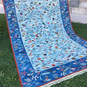 May include: A rectangular, hand-woven rug with a light blue central field and a dark blue border. The rug features a floral pattern with red, yellow, and white accents. The border has a red trim and white fringe.