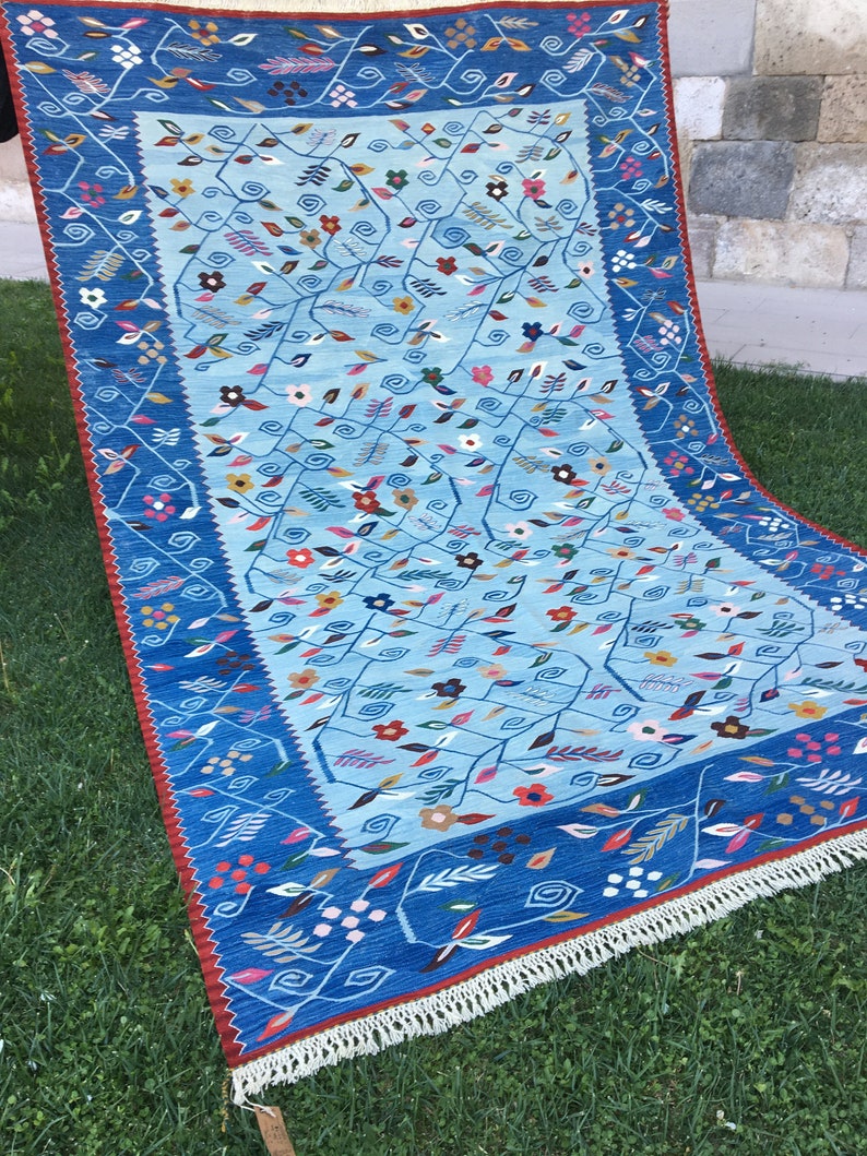 May include: A rectangular, hand-woven rug with a light blue central field and a dark blue border. The rug features a floral pattern with red, yellow, and white accents. The border has a red trim and white fringe.
