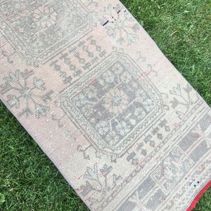 Op de afbeelding: Een vintage loper met een vervaagd roze en grijs kleurenschema. De loper heeft een geometrisch patroon met een centraal vierkant ontwerp. De loper heeft een rode franje rand.