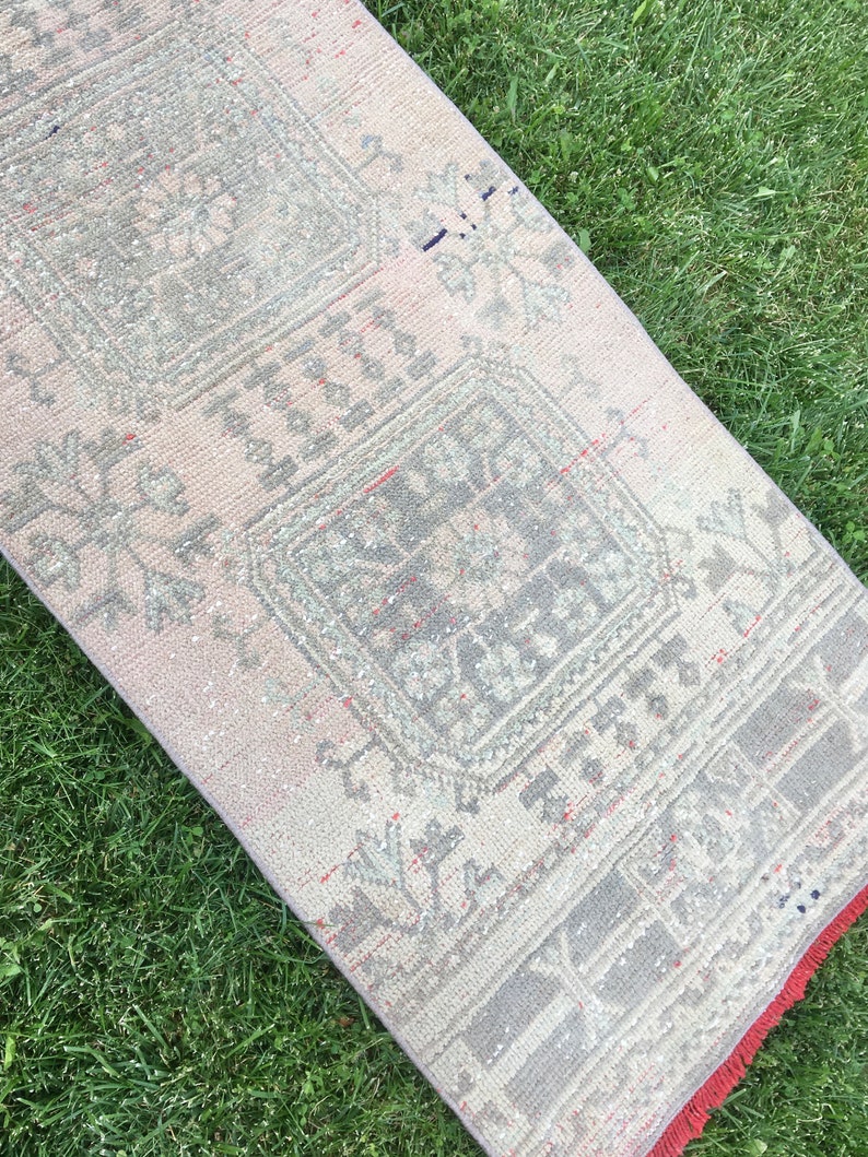 Op de afbeelding: Een vintage loper met een vervaagd roze en grijs kleurenschema. De loper heeft een geometrisch patroon met een centraal vierkant ontwerp. De loper heeft een rode franje rand.