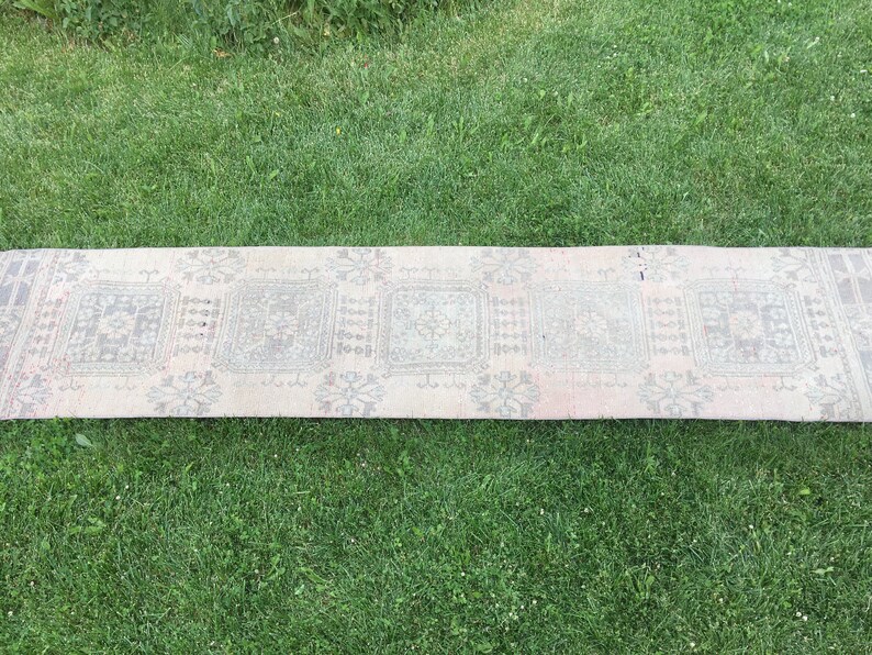 Op de afbeelding: Een lang, rechthoekig tapijt met een witte achtergrond en een herhalend patroon van grijze en bruine geometrische vormen. Het tapijt ligt op een groene grasmat.