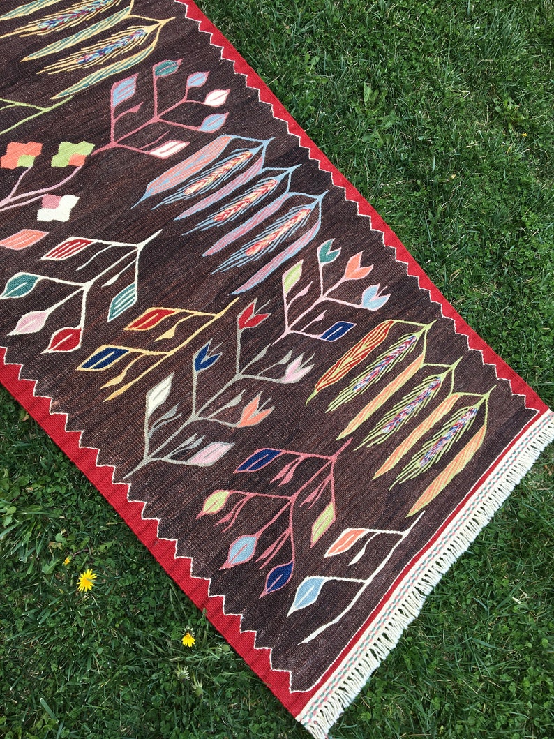Puede incluir: Una alfombra marr&oacute;n con un estampado floral colorido. La alfombra tiene un borde rojo y flecos blancos.