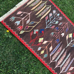 Puede incluir: Una alfombra marr&oacute;n y roja con un patr&oacute;n geom&eacute;trico de hojas estilizadas y espigas de trigo en tonos de azul, verde, amarillo, rosa y blanco.