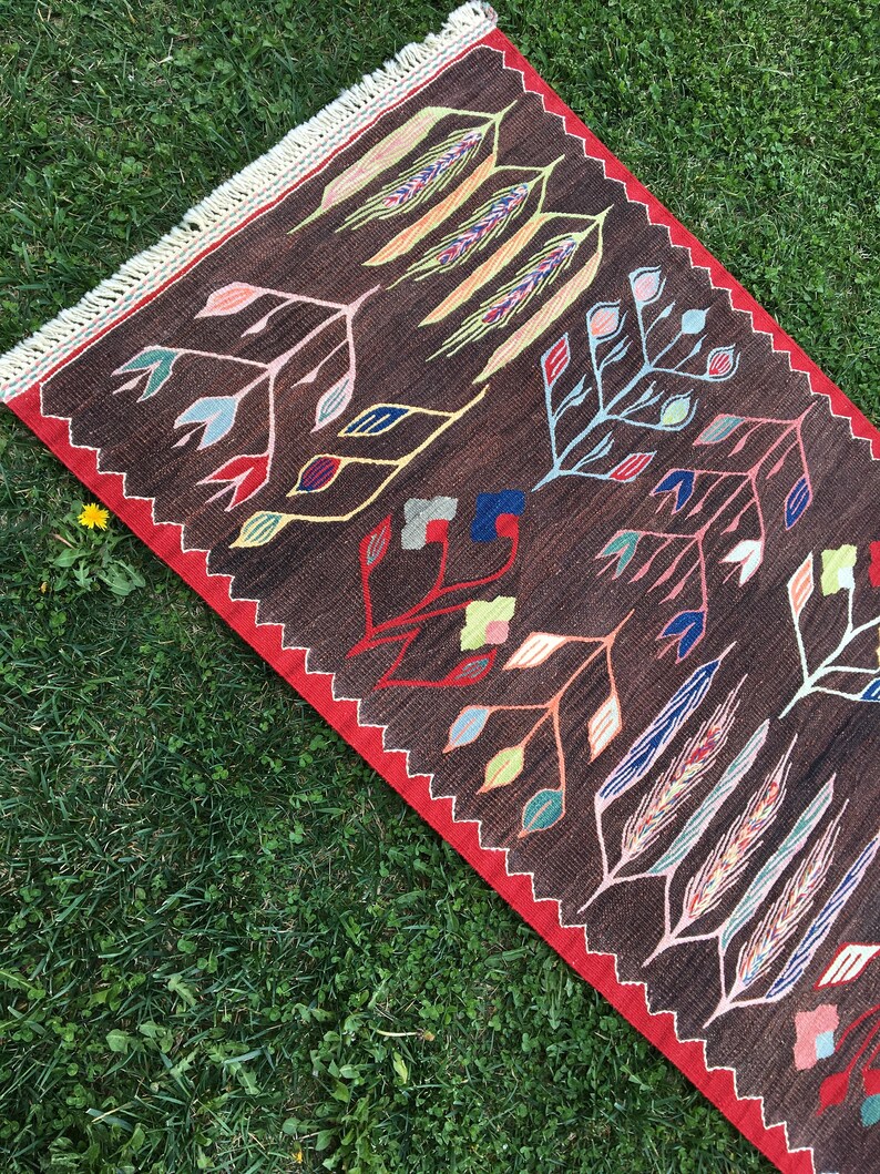 Puede incluir: Una alfombra marr&oacute;n y roja con un patr&oacute;n geom&eacute;trico de hojas estilizadas y espigas de trigo en tonos de azul, verde, amarillo, rosa y blanco.