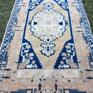 May include: A vintage rug with a beige background and a blue and white geometric pattern. The rug has a fringe border.