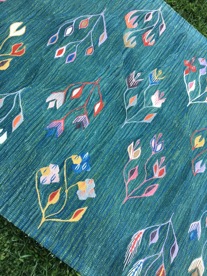 Puede incluir: Una alfombra azul verdosa con un patr&oacute;n tejido de flores y hojas de colores. La alfombra tiene una superficie texturizada y un brillo sutil.