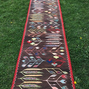 Puede incluir: Una alfombra larga y estrecha con fondo marr&oacute;n y un patr&oacute;n geom&eacute;trico colorido de hojas y flores estilizadas. La alfombra tiene un borde rojo y flecos en los extremos.