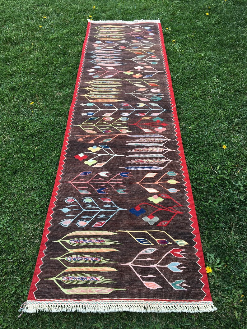Puede incluir: Una alfombra larga y estrecha con fondo marr&oacute;n y un patr&oacute;n geom&eacute;trico colorido de hojas y flores estilizadas. La alfombra tiene un borde rojo y flecos en los extremos.