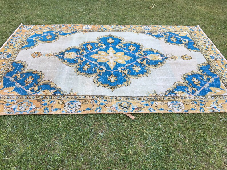 May include: A rectangular rug with a central medallion design. The rug features a light beige background with a large, ornate blue and yellow floral pattern. The border is a mix of yellow and blue, creating a vintage aesthetic.