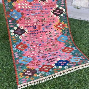 May include: A colourful rug with a floral pattern in shades of pink, red, green, blue, and yellow. The rug has a fringe border and is laid out on a green grassy surface.