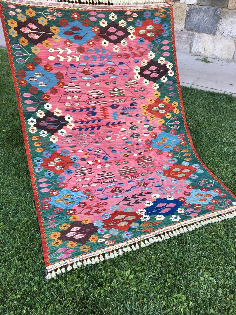 May include: A colourful rug with a floral pattern in shades of pink, red, green, blue, and yellow. The rug has a fringe border and is laid out on a green grassy surface.