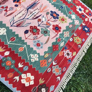May include: A colorful, floral-patterned rug with a pink central field. The rug features a repeating design of flowers and leaves in red, blue, green, and orange. The border is red with a light blue edge and white fringe.