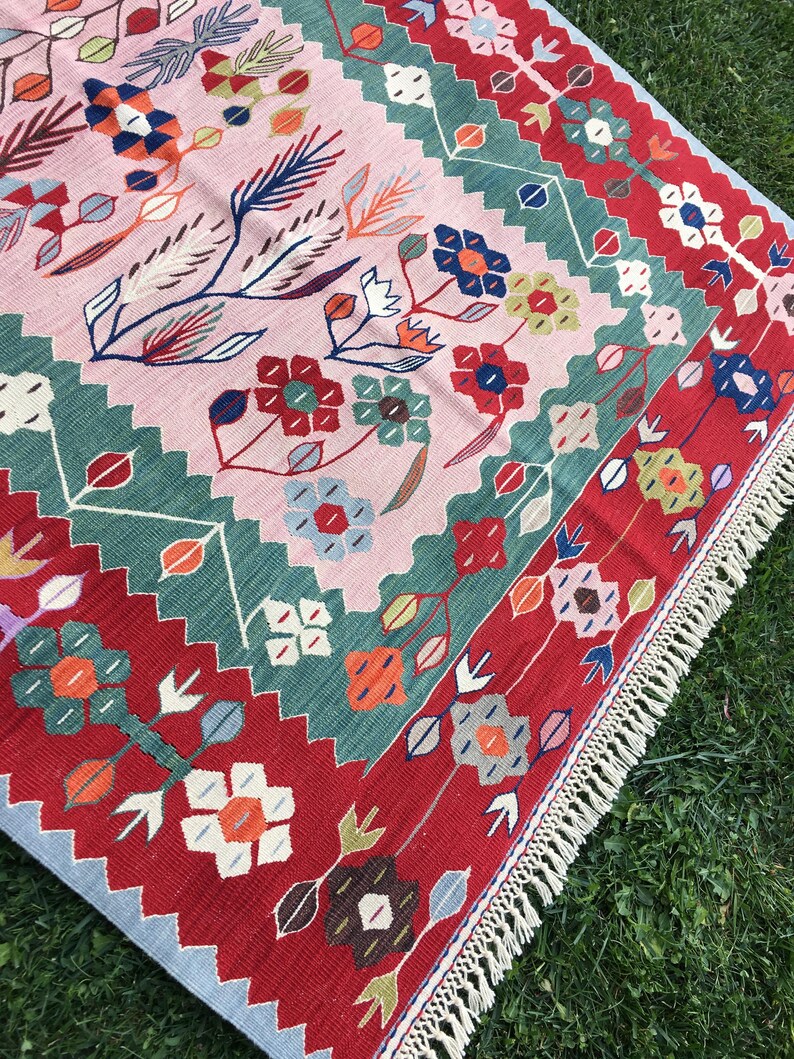 May include: A colorful, floral-patterned rug with a pink central field. The rug features a repeating design of flowers and leaves in red, blue, green, and orange. The border is red with a light blue edge and white fringe.