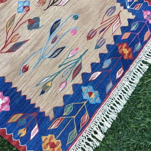May include: A beige rug with a blue and red border and a white fringe. The rug features a floral pattern with colorful flowers and leaves.