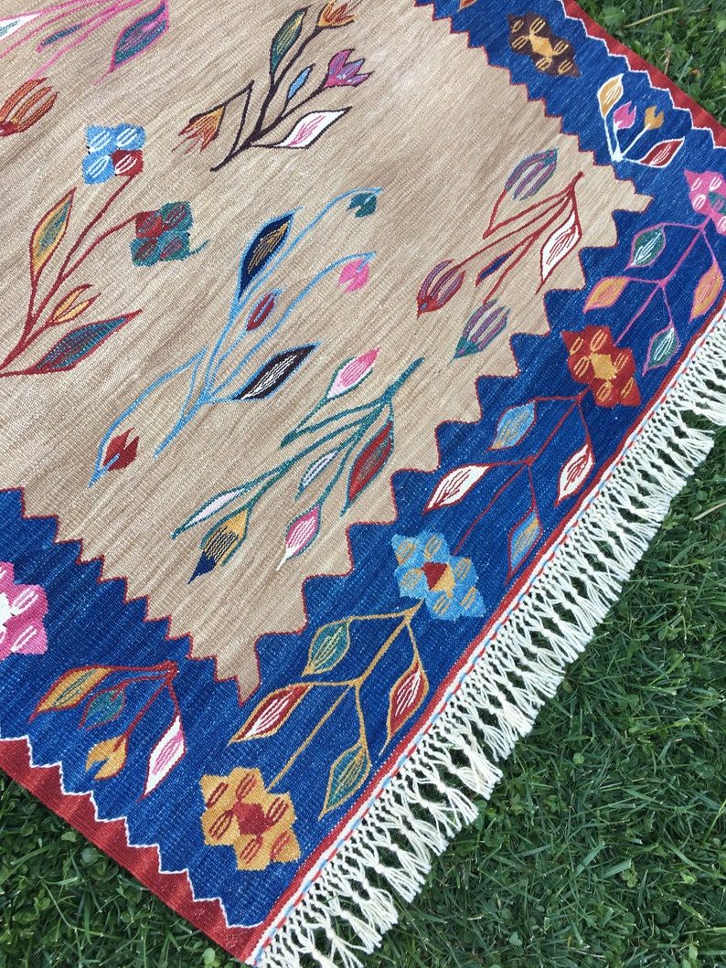 May include: A beige rug with a blue and red border and a white fringe. The rug features a floral pattern with colorful flowers and leaves.
