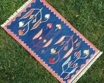 May include: A blue and red rug with a fringe border features a repeating pattern of stylized leaves and flowers in shades of yellow, orange, green, and blue.