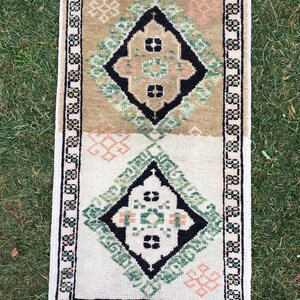 May include: A small, rectangular rug with a white background and a geometric pattern in shades of green, black, and pink. The rug has a fringed border.