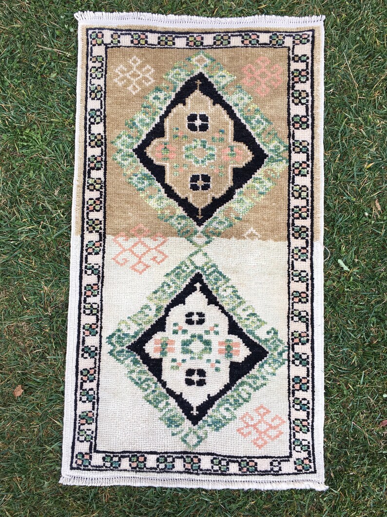May include: A small, rectangular rug with a white background and a geometric pattern in shades of green, black, and pink. The rug has a fringed border.