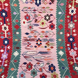 May include: A colorful, patterned rug with a central pink field. The rug features floral and geometric designs in red, green, blue, and white. The edges have a red border with white and blue floral motifs.