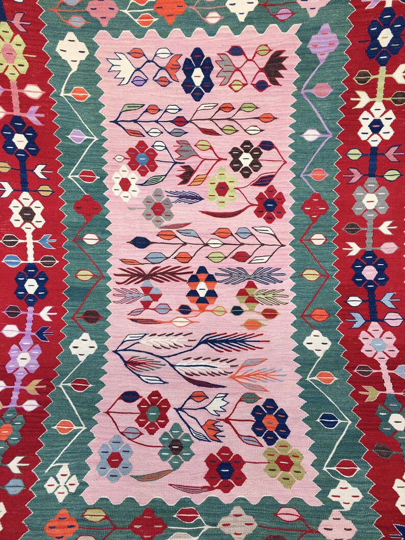 May include: A colorful, patterned rug with a central pink field. The rug features floral and geometric designs in red, green, blue, and white. The edges have a red border with white and blue floral motifs.