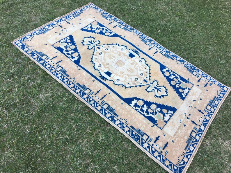 May include: A beige rug with a blue and white geometric pattern. The rug has a fringed edge and is laid out on a green grassy surface.
