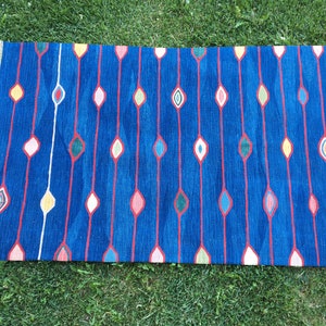 Puede incluir: Una alfombra azul con un patr&oacute;n geom&eacute;trico de l&iacute;neas rojas y formas de diamantes de colores. La alfombra tiene un borde de flecos.