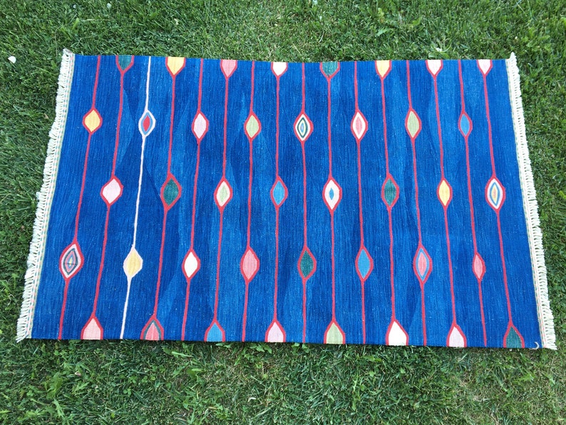 Puede incluir: Una alfombra azul con un patr&oacute;n geom&eacute;trico de l&iacute;neas rojas y formas de diamantes de colores. La alfombra tiene un borde de flecos.
