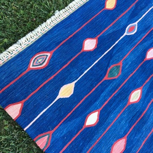 Puede incluir: Una alfombra azul con un patr&oacute;n geom&eacute;trico de diamantes de colores. La alfombra tiene un borde de flecos.