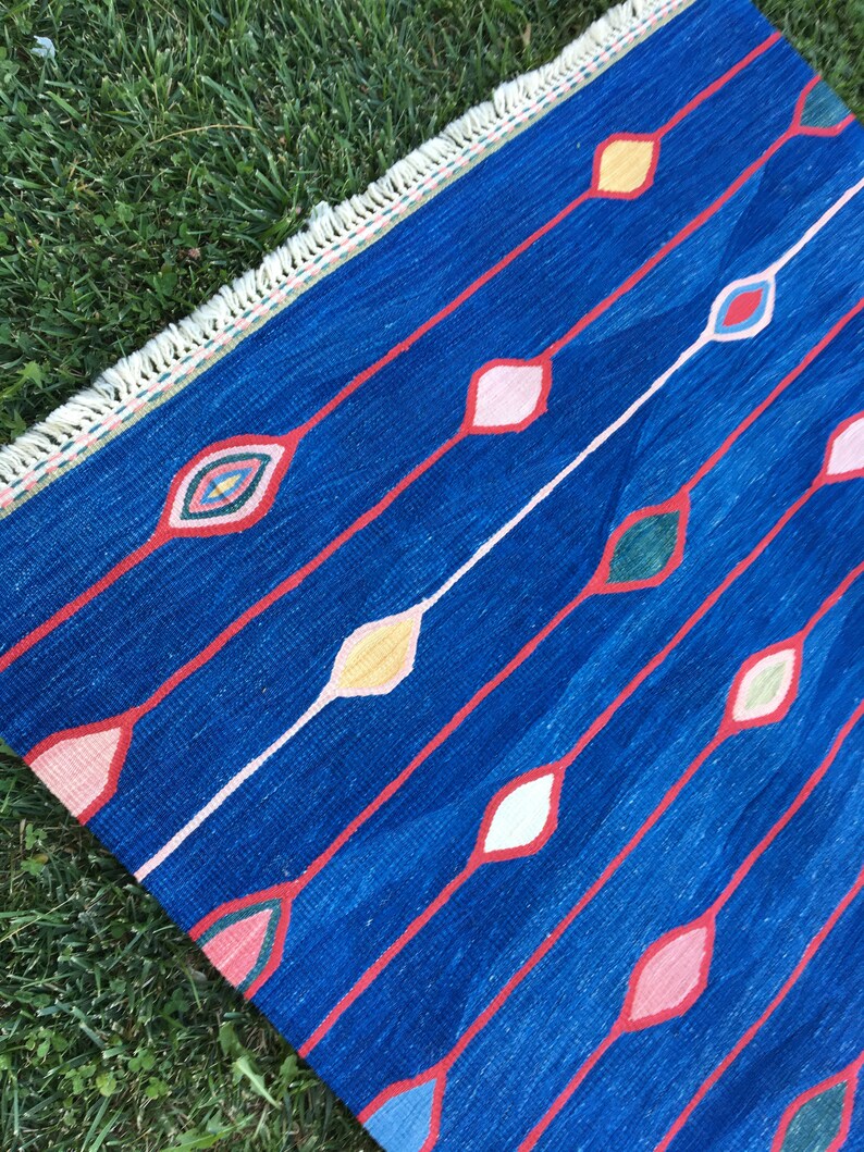 Puede incluir: Una alfombra azul con un patr&oacute;n geom&eacute;trico de diamantes de colores. La alfombra tiene un borde de flecos.