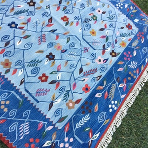 May include: A light blue rug with a floral pattern and a dark blue border. The rug features a repeating design of stylized flowers and leaves in shades of red, brown, and pink. The border has a geometric pattern and white fringe.