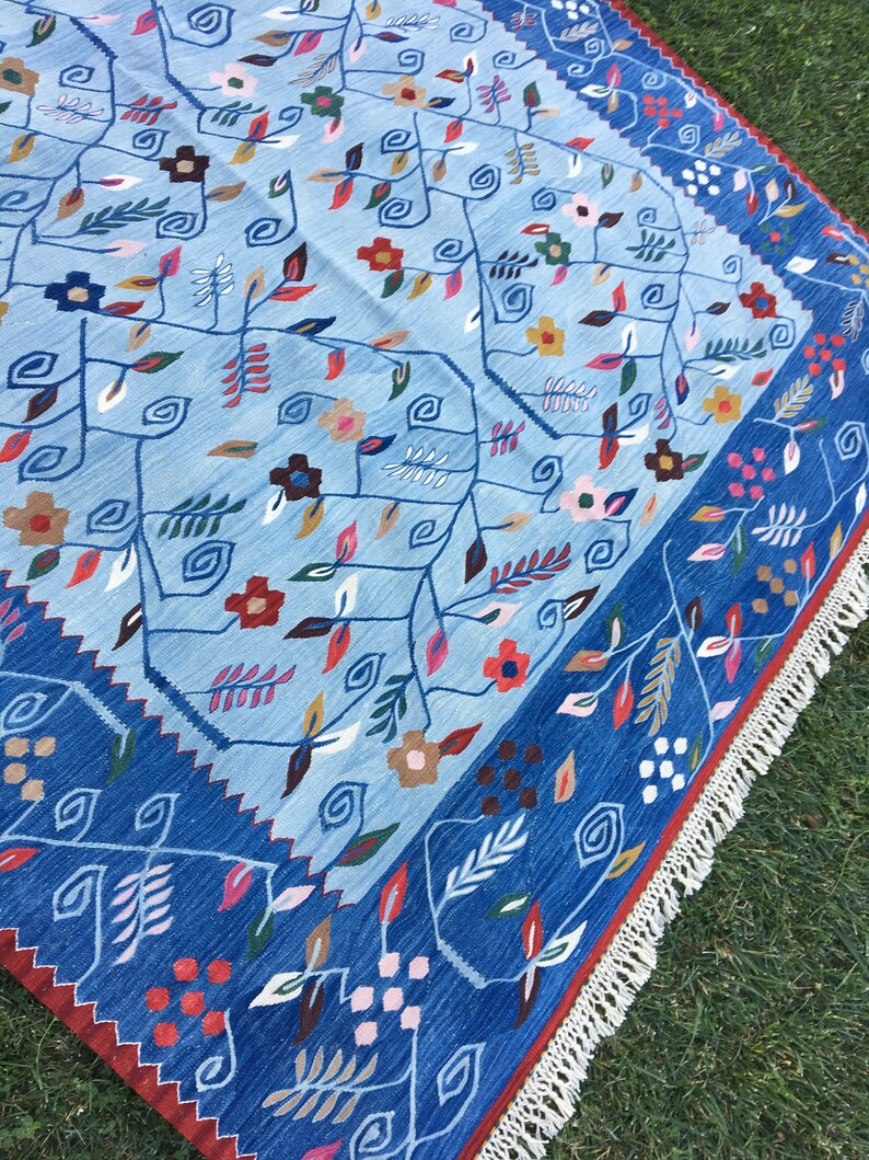 May include: A light blue rug with a floral pattern and a dark blue border. The rug features a repeating design of stylized flowers and leaves in shades of red, brown, and pink. The border has a geometric pattern and white fringe.
