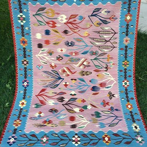 Op de afbeelding: Een kleurrijk tapijt met een bloemenpatroon in tinten roze, blauw, rood, groen en geel. Het tapijt heeft een blauwe rand met een geometrisch patroon en een rode rand met franjes.
