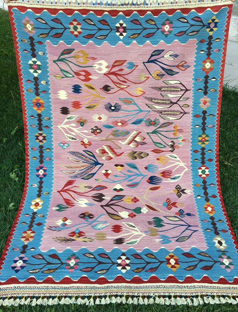 Op de afbeelding: Een kleurrijk tapijt met een bloemenpatroon in tinten roze, blauw, rood, groen en geel. Het tapijt heeft een blauwe rand met een geometrisch patroon en een rode rand met franjes.