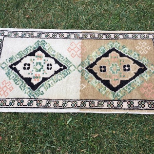 May include: A white and beige rug with a black and green geometric pattern. The rug has a fringed border.