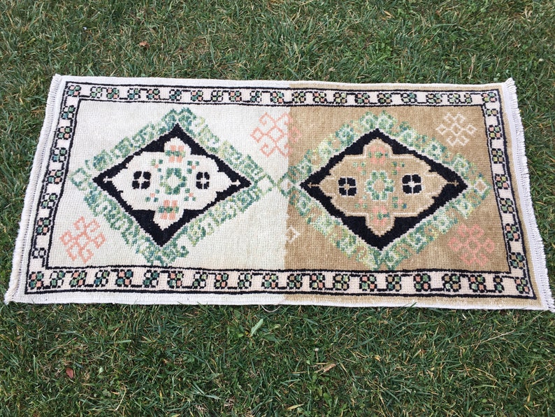 May include: A white and beige rug with a black and green geometric pattern. The rug has a fringed border.