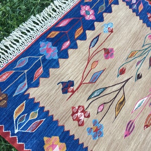 May include: A beige rug with a blue border and colorful floral designs. The rug has a fringe border.
