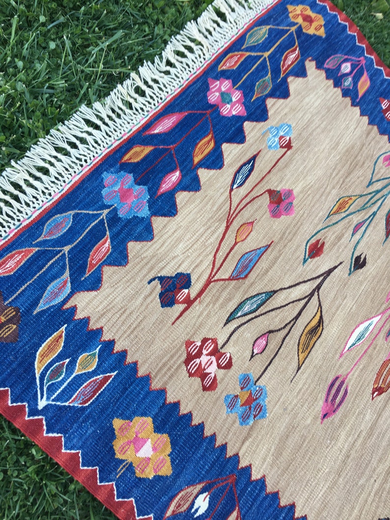 May include: A beige rug with a blue border and colorful floral designs. The rug has a fringe border.