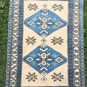 May include: A vintage rug with a cream background and a blue, white, and brown geometric pattern. The rug has a fringe border.