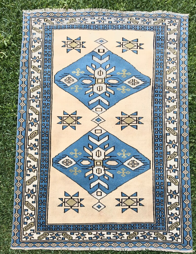 May include: A vintage rug with a cream background and a blue, white, and brown geometric pattern. The rug has a fringe border.
