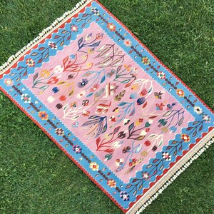 Op de afbeelding: Een roze en blauw tapijt met een bloemenpatroon. Het tapijt heeft een franje rand en is gemaakt van wol.