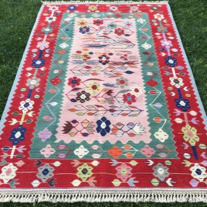 May include: A rectangular rug with a floral design. The rug features a red border with white fringe, a light pink center, and a green and blue floral pattern. The rug is made of woven material.