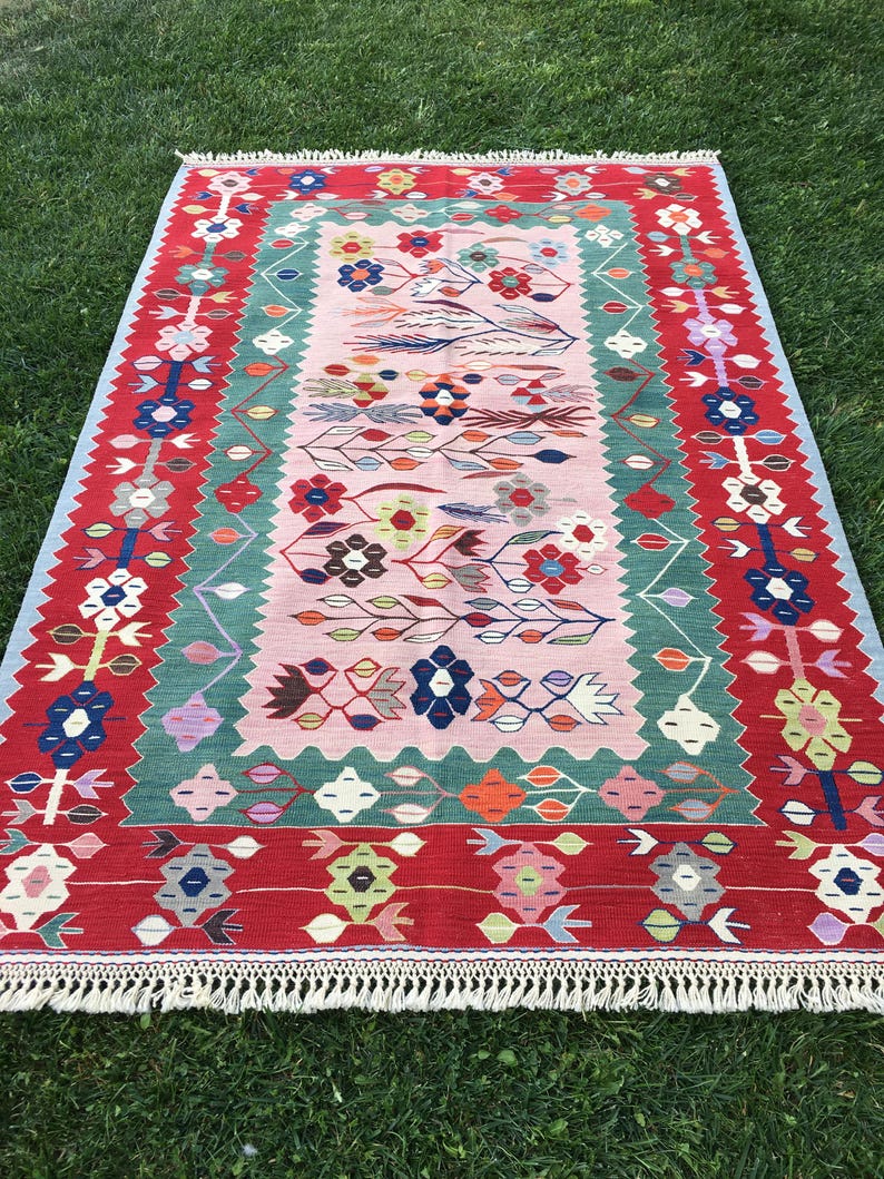 May include: A rectangular rug with a floral design. The rug features a red border with white fringe, a light pink center, and a green and blue floral pattern. The rug is made of woven material.