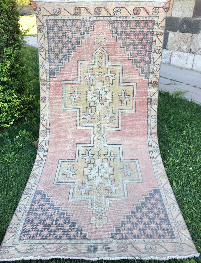 May include: A rectangular, vintage-style rug with a central geometric design in shades of peach, beige, and gold. The rug features a repeating pattern of floral and geometric shapes in the borders and corners, with accents of navy blue and gray. The rug is displayed outdoors on grass.
