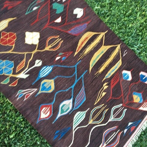 Puede incluir: Una alfombra marr&oacute;n con un patr&oacute;n geom&eacute;trico de hojas de colores y formas abstractas. La alfombra tiene un borde de flecos.