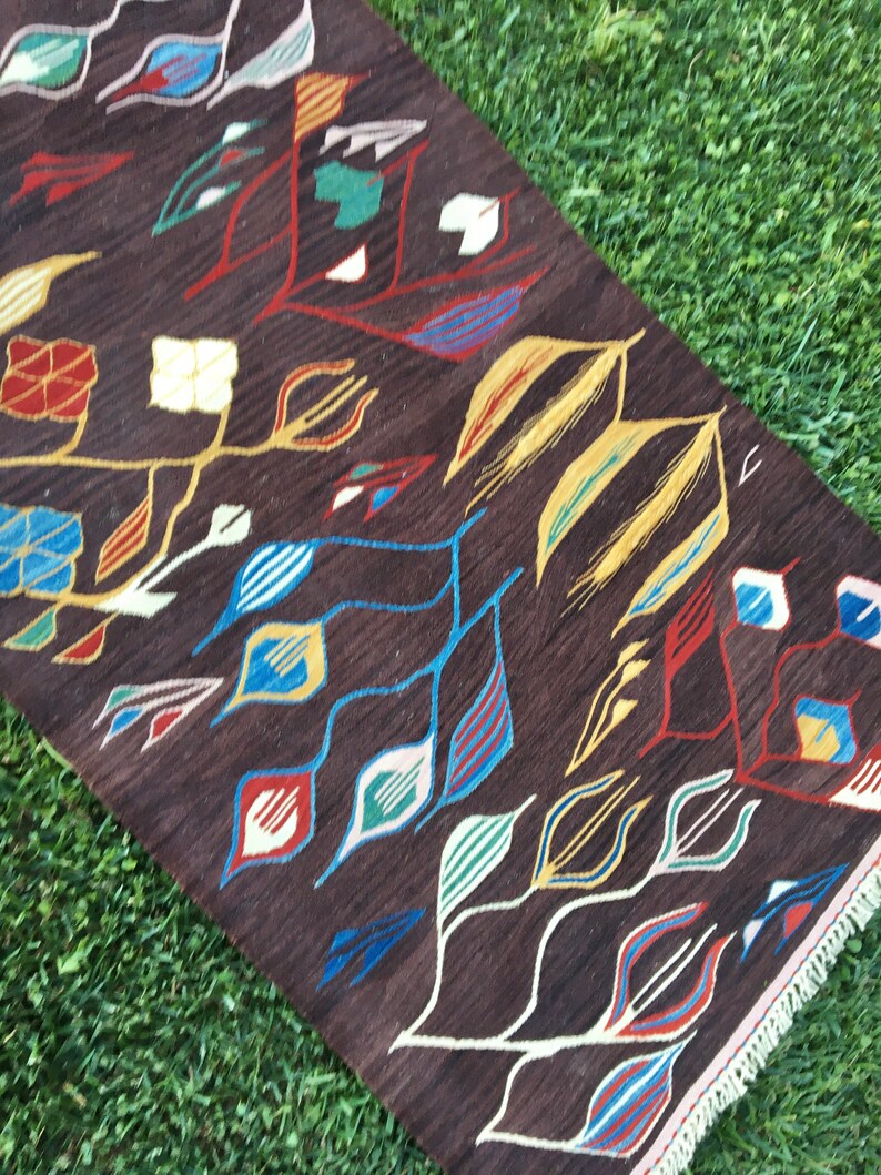 Puede incluir: Una alfombra marr&oacute;n con un patr&oacute;n geom&eacute;trico de hojas de colores y formas abstractas. La alfombra tiene un borde de flecos.