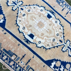 May include: A beige rug with a blue border and a white floral pattern in the center. The rug has a distressed look and is made of wool.