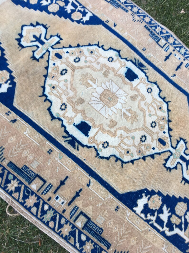 May include: A beige rug with a blue border and a white floral pattern in the center. The rug has a distressed look and is made of wool.