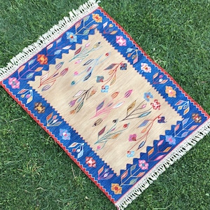 May include: A beige rug with a blue and red border. The rug has a floral pattern with pink, blue, and yellow flowers. The rug has a fringe border.